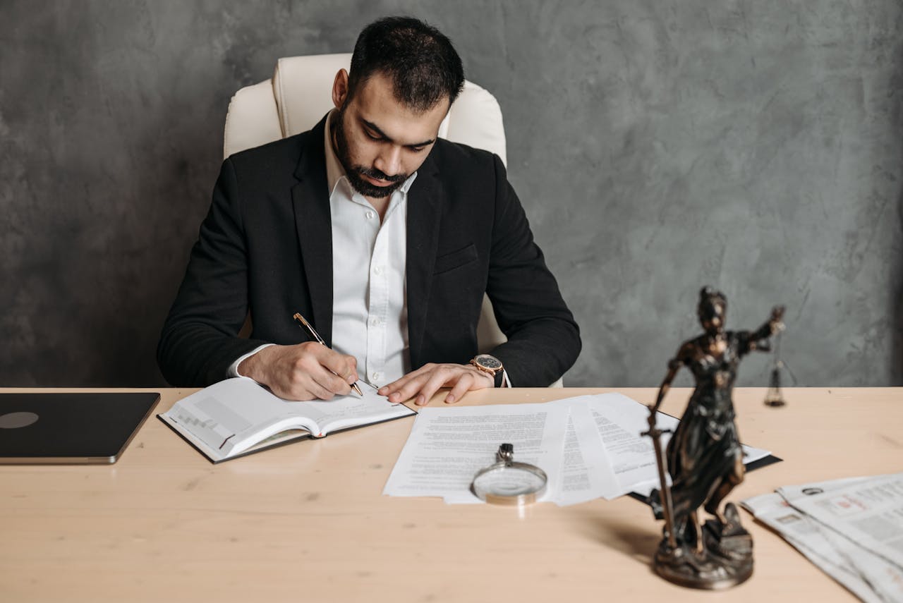 Focused lawyer in black suit at desk writing on documents in an office setting with legal statue.